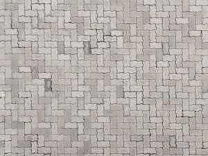 An arrangement of light gray interlocking paving stones forms a repetitive pattern. The pavers are rectangular with irregular, wavy edges, creating a uniform and structured surface.