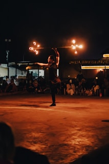A person is performing a fire dance on a stage at night, spinning a staff with flames. The audience is visible in the background, seated and watching attentively. The scene is illuminated by the orange glow of the fire, creating a striking contrast against the dark surroundings.