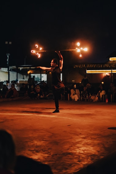 A person is performing a fire dance on a stage at night, spinning a staff with flames. The audience is visible in the background, seated and watching attentively. The scene is illuminated by the orange glow of the fire, creating a striking contrast against the dark surroundings.