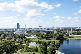 A sweeping view of an architectural structure with tensile roofs among lush greenery, with a small lake in the foreground and a cityscape in the distance. The sky is clear with a few scattered clouds.