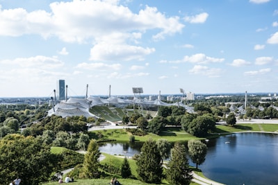 A sweeping view of an architectural structure with tensile roofs among lush greenery, with a small lake in the foreground and a cityscape in the distance. The sky is clear with a few scattered clouds.