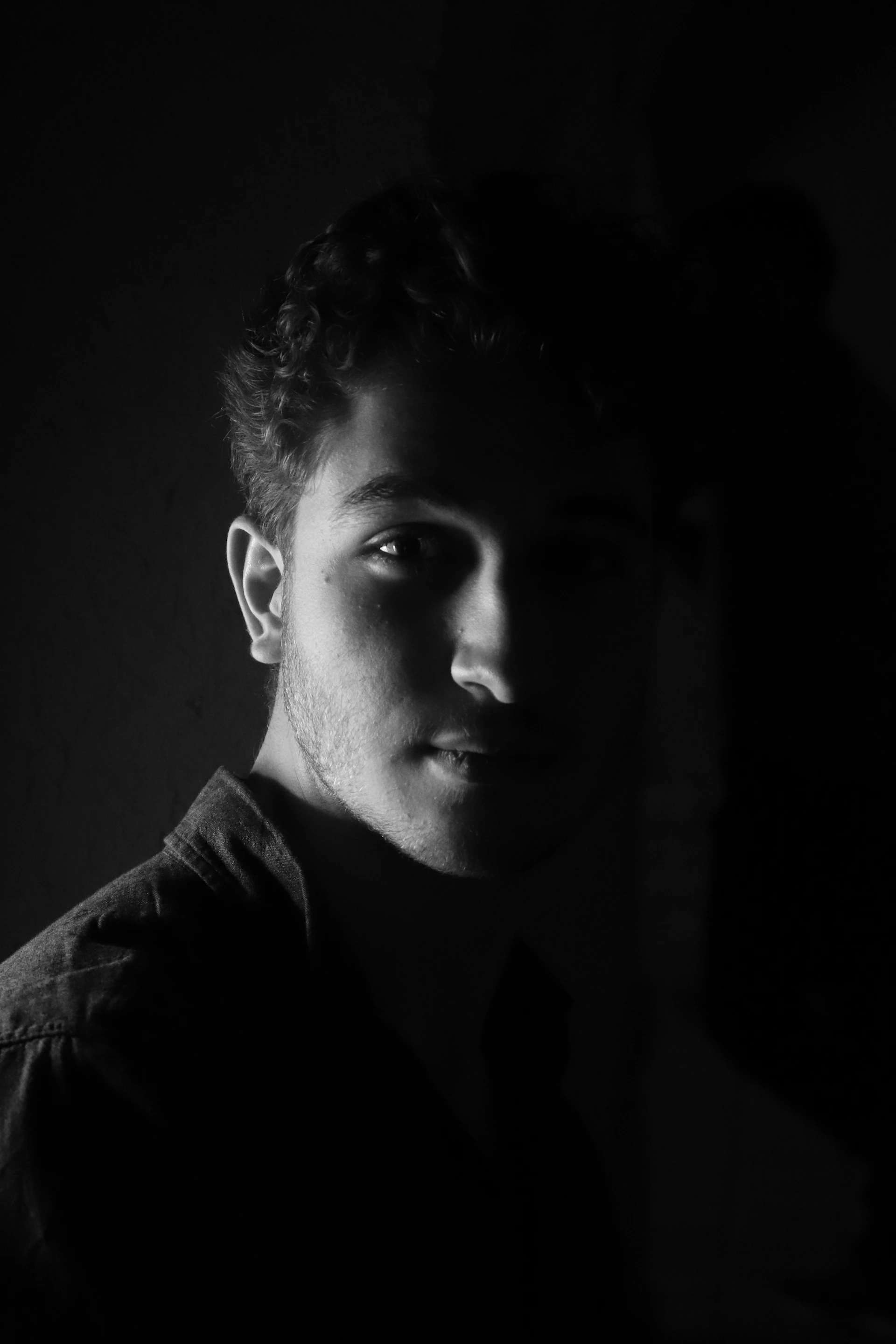 A moody black-and-white portrait of Profit Elijah standing against a textured brick wall in Brooklyn at night, with subtle fog curling around him and soft gold light highlighting his profile.