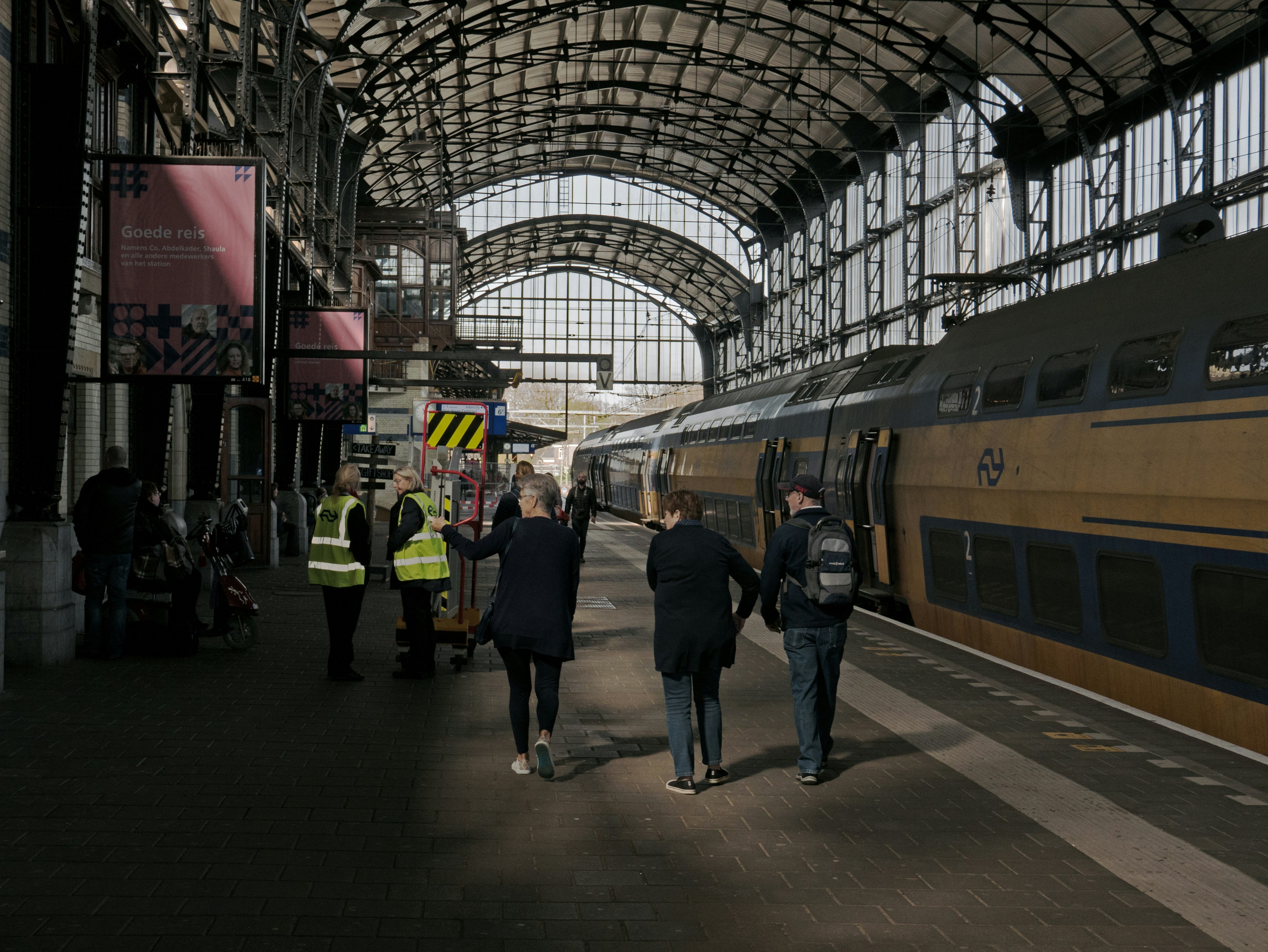 Train conductors and travellers are walking and standing at the platform, close to the waiting yellow Intercity train, direction Haarlem. This dark location is part of Central station Amsterdam, with the large old smoke cap, built c. 1880 // Foto van mensen die lopen in de oude stationshal met zijn hoge dak en bogen, op het treinstation van Haarlem - oude architectuur met zijn bijzondere metalen dakconstructie. De gele Intercity is net aangekomen op het perron. Foto openbaar vervoer van Fons Heijnsbroek - straatfotografie uit Nederland in hoge resolutie en rechtenvrije afbeelding, CC0.