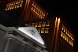 A nighttime view of a building with illuminated windows and an architectural pediment featuring a Star of David. The structure displays classical elements with columns and detailed molding, contrasted against a modern backdrop of lit windows.