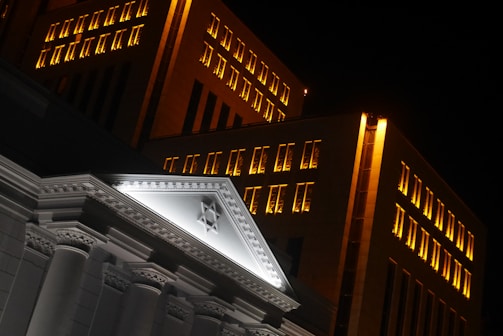 A nighttime view of a building with illuminated windows and an architectural pediment featuring a Star of David. The structure displays classical elements with columns and detailed molding, contrasted against a modern backdrop of lit windows.