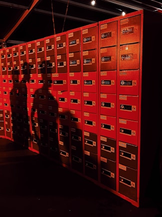 A wall of red lockers is illuminated by warm lighting, casting shadows of two people against the surface. The lockers are labeled with numbers and feature combination locks.