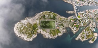 An aerial view of a small island featuring a football field at its center, surrounded by rocky terrain and greenery. The island is located in a body of water with a nearby coastal town visible towards the upper right corner. Narrow roads connect structures on the island, with residential houses visible in the coastal town.
