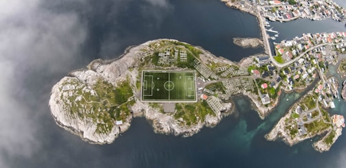 An aerial view of a small island featuring a football field at its center, surrounded by rocky terrain and greenery. The island is located in a body of water with a nearby coastal town visible towards the upper right corner. Narrow roads connect structures on the island, with residential houses visible in the coastal town.
