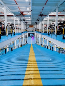 A multi-level car parking structure with blue floors and a central yellow path. Several cars are parked on both sides of the levels. Bright overhead lighting illuminates the space, which features white walls and red pipes along the ceiling.