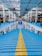 A multi-level car parking structure with blue floors and a central yellow path. Several cars are parked on both sides of the levels. Bright overhead lighting illuminates the space, which features white walls and red pipes along the ceiling.