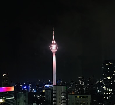 A tall telecommunications tower dominates the city skyline at night, brightly illuminated against a dark sky. Surrounding buildings display various colors in their lights, contributing to a vibrant urban scene.
