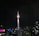 A tall telecommunications tower dominates the city skyline at night, brightly illuminated against a dark sky. Surrounding buildings display various colors in their lights, contributing to a vibrant urban scene.