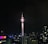 A tall telecommunications tower dominates the city skyline at night, brightly illuminated against a dark sky. Surrounding buildings display various colors in their lights, contributing to a vibrant urban scene.