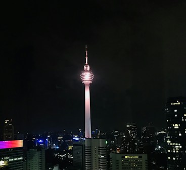 Uma torre de telecomunicações domina o horizonte urbano à noite, iluminada contra um céu escuro.