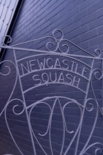 A decorative wrought iron sign displaying the words 'Newcastle Squash' is positioned against a painted brick wall. The intricate design includes swirls and curves, adding an artistic touch to the functional sign.