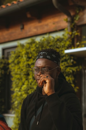 A person wearing glasses and a black bandana stands outdoors. They are dressed in a black hoodie and appear to be in a thoughtful pose. The background features green foliage and a wooden building, suggesting a natural and calm environment.