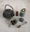 A textured, dark metal teapot with a curved handle sits alongside a small green dish filled with dried tea leaves. A lit green candle is positioned next to a multi-colored stone, a polished brown crystal point, and a decorative gold spoon with an intricate design.