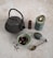 A textured, dark metal teapot with a curved handle sits alongside a small green dish filled with dried tea leaves. A lit green candle is positioned next to a multi-colored stone, a polished brown crystal point, and a decorative gold spoon with an intricate design.