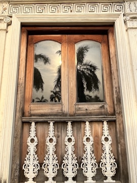 A wooden window with two arched panes reflecting palm trees and the sun. The window is framed by intricate white carvings, with ornate white metalwork railing below. The architectural style is detailed, featuring decorative patterns above the window.