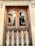 A wooden window with two arched panes reflecting palm trees and the sun. The window is framed by intricate white carvings, with ornate white metalwork railing below. The architectural style is detailed, featuring decorative patterns above the window.