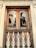 A wooden window with two arched panes reflecting palm trees and the sun. The window is framed by intricate white carvings, with ornate white metalwork railing below. The architectural style is detailed, featuring decorative patterns above the window.