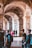 A group of people walking through an ancient stone corridor with high, vaulted ceilings and arches. The interior is illuminated with natural light, highlighting the texture and details of the stonework. Some individuals carry backpacks, and there is a sense of movement and exploration.