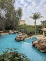 A serene outdoor scene featuring a lagoon-style swimming pool surrounded by lush greenery, including various trees and bushes. Large rocks are integrated into the pool's design, and a small cascade feature adds movement to the water. The sky is partly cloudy, adding a sense of calm to the environment.