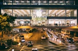 A large, busy Apple store occupies an upper floor of a modern building with glass windows. People are visible inside the store, which is bustling with activity. Outside, a street is filled with traffic, including cars and a double-decker bus, all moving beneath the brightly lit store.