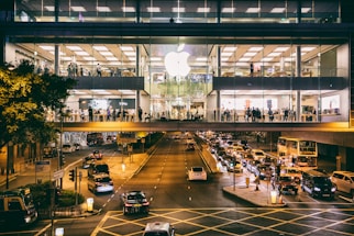 A large, busy Apple store occupies an upper floor of a modern building with glass windows. People are visible inside the store, which is bustling with activity. Outside, a street is filled with traffic, including cars and a double-decker bus, all moving beneath the brightly lit store.