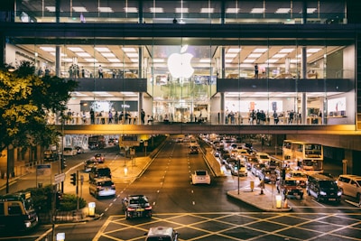 A large, busy Apple store occupies an upper floor of a modern building with glass windows. People are visible inside the store, which is bustling with activity. Outside, a street is filled with traffic, including cars and a double-decker bus, all moving beneath the brightly lit store.