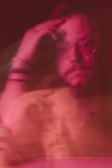 A blurred, artistic portrait of a person with a contemplative expression. The colors are predominantly pink and red, and the double exposure effect creates a sense of motion and ambiguity.