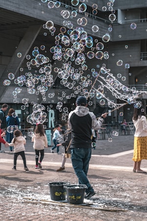 Numerous soap bubbles floating in the air with a street performer using sticks to create them. Children are joyfully playing and running around, attempting to catch the bubbles. The scene takes place in an urban setting with a concrete building in the background.