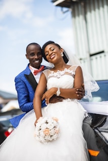 A joyful couple dressed in wedding attire, with the groom in a blue suit and red bow tie, and the bride in a white dress, holding a bouquet of flowers.