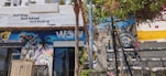 A vibrant street scene featuring a surf shop with signage offering surf school, guiding, and yoga. The exterior wall is adorned with colorful murals, including an eye-catching image resembling the Statue of Liberty and a spray can. Lush green foliage and bright flowering plants partially cover the upper part of the wall, adding a natural element. The atmosphere seems lively and artistic.
