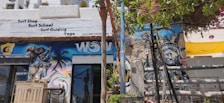 A vibrant street scene featuring a surf shop with signage offering surf school, guiding, and yoga. The exterior wall is adorned with colorful murals, including an eye-catching image resembling the Statue of Liberty and a spray can. Lush green foliage and bright flowering plants partially cover the upper part of the wall, adding a natural element. The atmosphere seems lively and artistic.