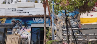 A vibrant street scene featuring a surf shop with signage offering surf school, guiding, and yoga. The exterior wall is adorned with colorful murals, including an eye-catching image resembling the Statue of Liberty and a spray can. Lush green foliage and bright flowering plants partially cover the upper part of the wall, adding a natural element. The atmosphere seems lively and artistic.