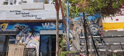 A vibrant street scene featuring a surf shop with signage offering surf school, guiding, and yoga. The exterior wall is adorned with colorful murals, including an eye-catching image resembling the Statue of Liberty and a spray can. Lush green foliage and bright flowering plants partially cover the upper part of the wall, adding a natural element. The atmosphere seems lively and artistic.