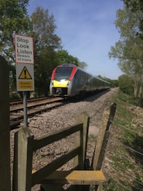 A train is traveling on railway tracks surrounded by greenery. A safety sign is prominently displayed in the foreground, advising caution around trains. The tracks stretch into the distance, flanked by trees and shrubs under a clear blue sky.