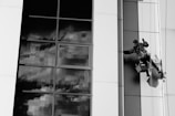 A person is suspended on a rope cleaning the windows of a modern building with large glass panels. The reflection in the glass shows a cloudy sky, creating a dramatic contrast against the smooth, sleek architecture. The worker is equipped with a harness and is holding cleaning tools.