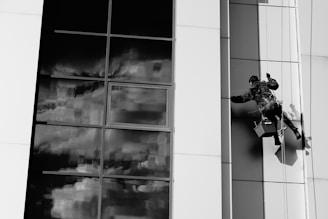 A person is suspended on a rope cleaning the windows of a modern building with large glass panels. The reflection in the glass shows a cloudy sky, creating a dramatic contrast against the smooth, sleek architecture. The worker is equipped with a harness and is holding cleaning tools.