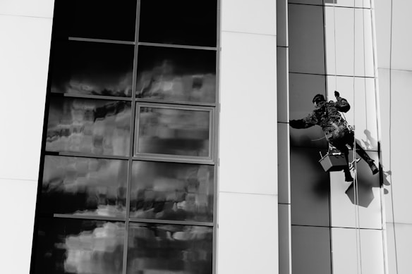 A person is suspended on a rope cleaning the windows of a modern building with large glass panels. The reflection in the glass shows a cloudy sky, creating a dramatic contrast against the smooth, sleek architecture. The worker is equipped with a harness and is holding cleaning tools.