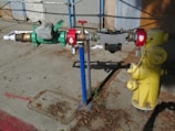A yellow fire hydrant is connected to a complex assembly of valves and pipes, including a prominent green mechanical device with gauges. The equipment is mounted on a concrete sidewalk, with scattered dried leaves and debris around it. A blue pole supports part of the assembly, and there are red connectors at various points.
