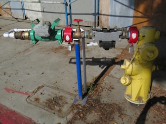 A yellow fire hydrant is connected to a complex assembly of valves and pipes, including a prominent green mechanical device with gauges. The equipment is mounted on a concrete sidewalk, with scattered dried leaves and debris around it. A blue pole supports part of the assembly, and there are red connectors at various points.