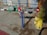 A yellow fire hydrant is connected to a complex assembly of valves and pipes, including a prominent green mechanical device with gauges. The equipment is mounted on a concrete sidewalk, with scattered dried leaves and debris around it. A blue pole supports part of the assembly, and there are red connectors at various points.