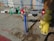 A yellow fire hydrant is connected to a complex assembly of valves and pipes, including a prominent green mechanical device with gauges. The equipment is mounted on a concrete sidewalk, with scattered dried leaves and debris around it. A blue pole supports part of the assembly, and there are red connectors at various points.
