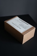 A cardboard box is placed on a dark surface. The box has a label on top with text related to personal delivery. Soft lighting creates subtle shadows around the box, emphasizing its shape and texture.