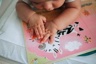 A baby's hands are gently placed on a colorful storybook. The book features illustrations of animals, including a zebra, with a pink and green backdrop. The infant is focused on the images, providing a sense of curiosity and learning.