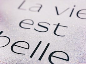 A close-up view of text on a sparkling background with the phrase 'La vie est belle' written in elegant black font. The surface appears to be glittery, creating a shimmering effect around the letters.