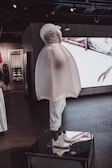A mannequin dressed in a unique, inflated outerwear piece and white sneakers. The figure is displayed on a platform inside a modern store with dark floors and ambient lighting. Behind it, there are shelves and a large screen showing abstract visuals.