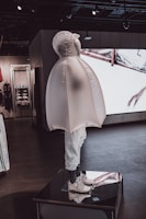 A mannequin dressed in a unique, inflated outerwear piece and white sneakers. The figure is displayed on a platform inside a modern store with dark floors and ambient lighting. Behind it, there are shelves and a large screen showing abstract visuals.